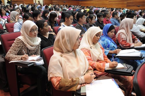 Students in a very full lecture theatre. Many of them are wearing hijabs. They are looking studious and either looking forwards or they are writing notes.